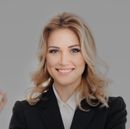Woman in a black suit smiles; blonde hair, white shirt, light background.